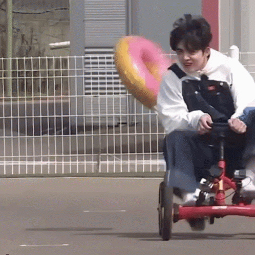 A Young Man Is Riding A Tricycle With A Pink Donut In The Back . GIF