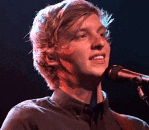 A Young Man Is Singing Into A Microphone While Playing A Guitar On Stage . GIF