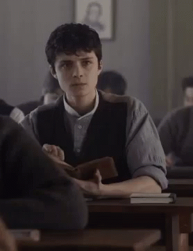 A Young Man Is Sitting At A Desk In A Classroom Holding A Book GIF