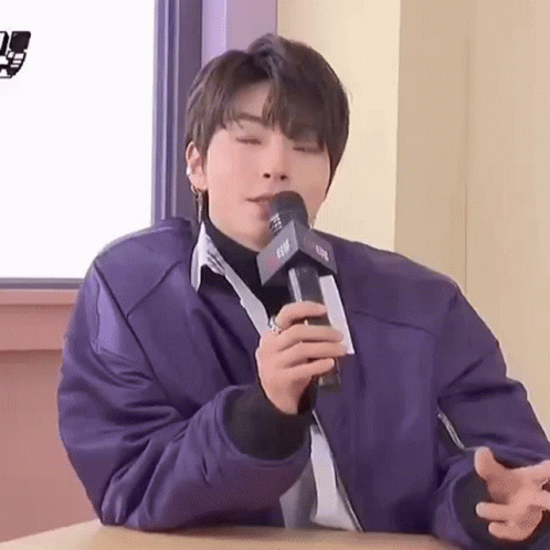 A Young Man Is Sitting At A Table Holding A Microphone In His Hand . GIF