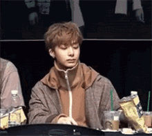 A Young Man Is Sitting At A Table With A Drink In His Hand And A Bag Of Chips . GIF