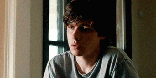 A Young Man Is Sitting In Front Of A Window Wearing A Grey Shirt . GIF