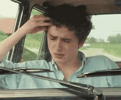A Young Man Is Sitting In The Driver 'S Seat Of A Car Holding His Head . GIF
