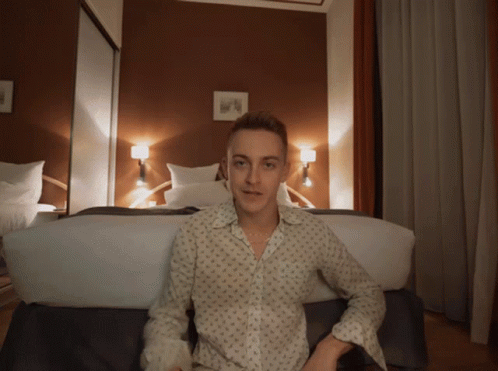 A Young Man Is Sitting On A Bed In A Bedroom GIF