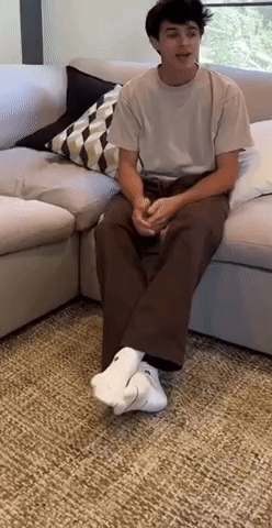 A Young Man Is Sitting On A Couch Wearing White Socks And A White Shirt . GIF