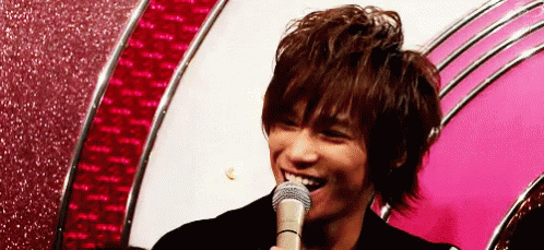 A Young Man Is Smiling While Holding A Microphone In Front Of A Pink Wall . GIF