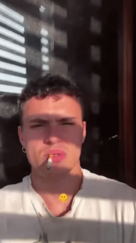 A Young Man Is Smoking A Cigarette In Front Of A Window While Wearing A Smiley Face Necklace . GIF