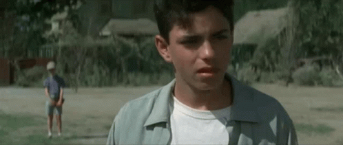 A Young Man Is Standing In A Field Looking At The Camera While Another Young Man Stands In The Background . GIF