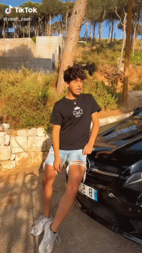 A Young Man Is Standing In Front Of A Black Car . GIF