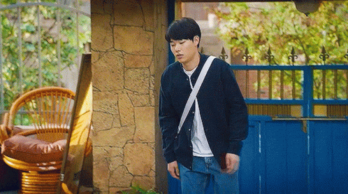 A Young Man Is Standing In Front Of A Blue Fence And A Wicker Chair . GIF