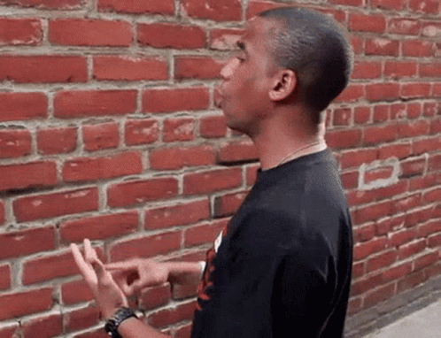 A Young Man Is Standing In Front Of A Red Brick Wall . GIF