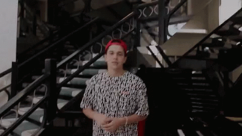A Young Man Is Standing In Front Of A Set Of Stairs Wearing A Red Hat And A Black And White Shirt . GIF