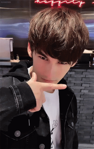 A Young Man Making A Peace Sign With His Finger In Front Of A Neon Sign That Says Happy GIF