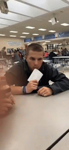 A Young Man Sitting At A Table In A Cafeteria Holding A Piece Of Paper GIF