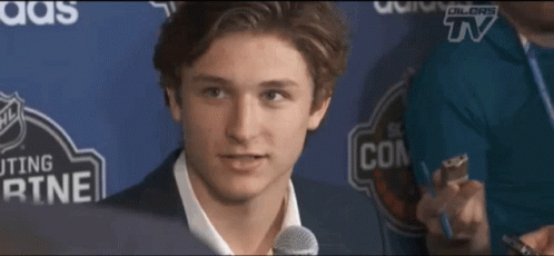 A Young Man Speaking Into A Microphone In Front Of A Sign That Says ' Dillors Tv ' On It GIF