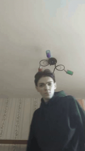 A Young Man Stands In Front Of A Ceiling Light With A Lamp On Top Of His Head GIF