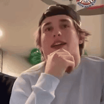 A Young Man Wearing A Baseball Cap And A White Sweater Is Sitting In A Living Room With His Hands On His Chin . GIF