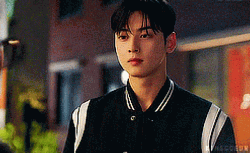 A Young Man Wearing A Black And White Baseball Jacket Is Looking At The Camera . GIF