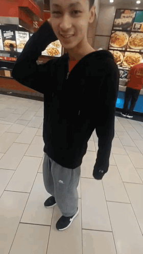 A Young Man Wearing A Black Hoodie And Grey Sweatpants Is Standing In Front Of A Fast Food Restaurant GIF