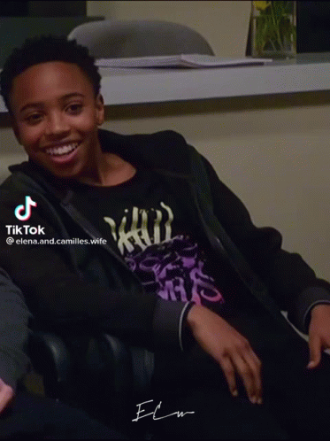 A Young Man Wearing A Black Jacket And A Purple Shirt Is Smiling And Sitting In A Chair GIF