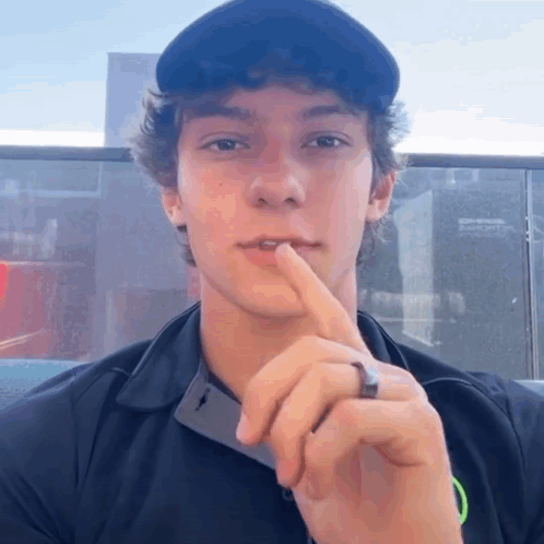 A Young Man Wearing A Black Shirt And A Black Hat Holds His Finger To His Lips GIF