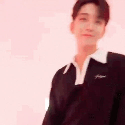 A Young Man Wearing A Black Sweater And A White Collar Is Standing In Front Of A White Wall . GIF