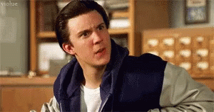 A Young Man Wearing A Blue And Gray Jacket Is Sitting In A Library . GIF
