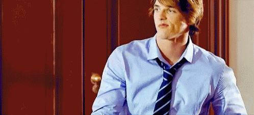 A Young Man Wearing A Blue Shirt And Tie Is Standing In Front Of A Door . GIF