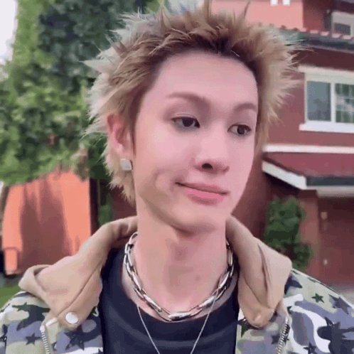 A Young Man Wearing A Camouflage Jacket And A Necklace Looks At The Camera GIF