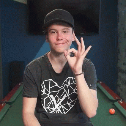 A Young Man Wearing A Hat And A Black Shirt Is Sitting In Front Of A Pool Table Giving An Ok Sign . GIF