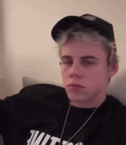 A Young Man Wearing A Hat And A Black Shirt Is Sitting On A Couch . GIF