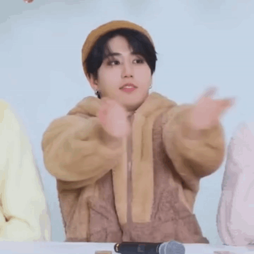 A Young Man Wearing A Hat And A Brown Jacket Is Sitting At A Table . GIF