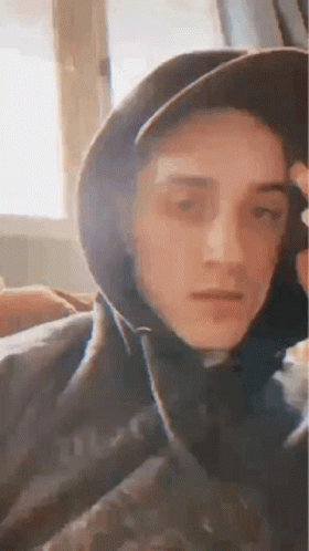 A Young Man Wearing A Hat And A Hoodie Is Sitting In Front Of A Window . GIF