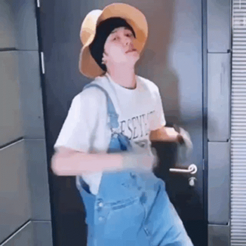 A Young Man Wearing A Hat And Overalls Is Dancing In Front Of A Door . GIF