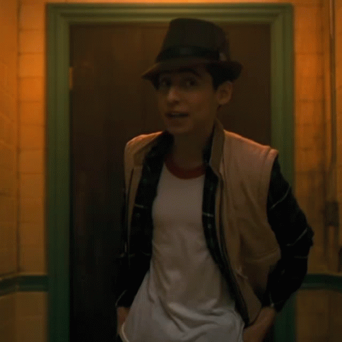 A Young Man Wearing A Hat And Vest Is Standing In A Doorway GIF