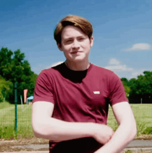 A Young Man Wearing A Maroon T-Shirt Is Standing In Front Of A Field GIF