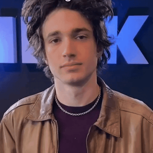 A Young Man Wearing A Necklace And A Leather Jacket Looks At The Camera GIF