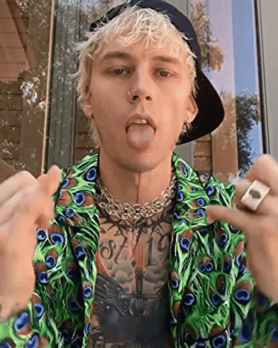 A Young Man Wearing A Peacock Feather Shirt And A Hat Is Sticking His Tongue Out . GIF
