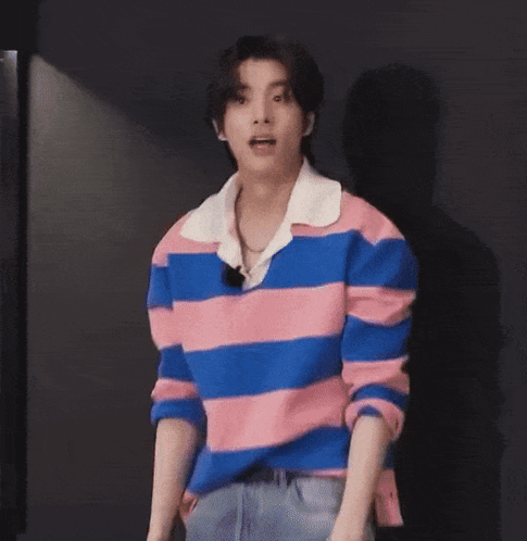 A Young Man Wearing A Pink And Blue Striped Shirt Is Standing In Front Of A Black Wall With His Mouth Open . GIF