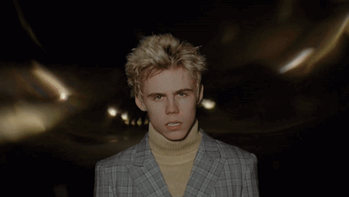 A Young Man Wearing A Plaid Jacket And A Yellow Turtleneck Looks At The Camera GIF