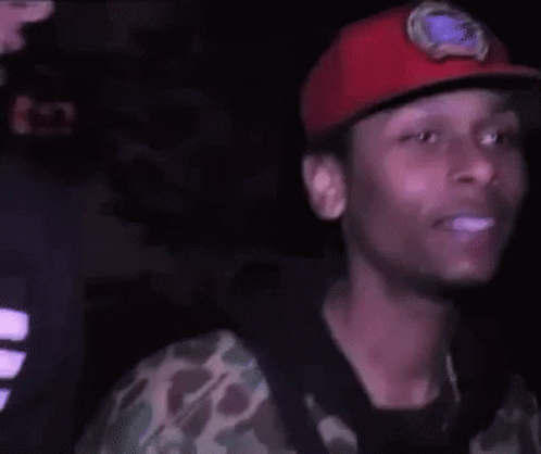 A Young Man Wearing A Red Hat And A Camouflage Shirt Is Smiling In The Dark . GIF