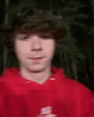 A Young Man Wearing A Red Hoodie Is Standing In Front Of A Tree . GIF