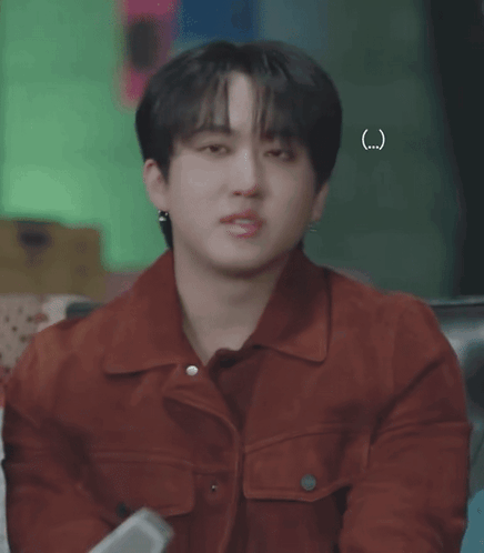 A Young Man Wearing A Red Jacket And Earrings Is Sitting On A Couch . GIF