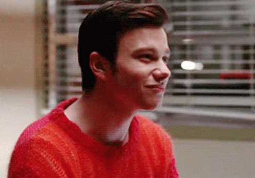 A Young Man Wearing A Red Sweater Is Smiling While Sitting In Front Of A Window . GIF
