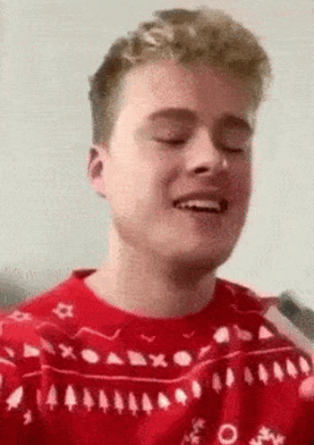A Young Man Wearing A Red Sweater With Trees On It Is Smiling With His Eyes Closed . GIF