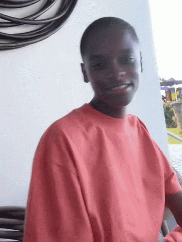 A Young Man Wearing A Red Sweatshirt Is Smiling While Sitting On A Balcony . GIF