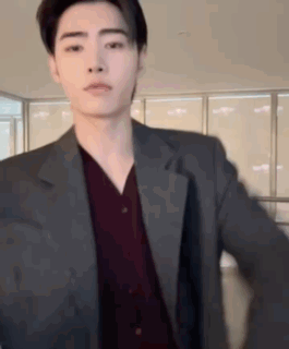 A Young Man Wearing A Suit And A Red Shirt Is Standing In Front Of A Window . GIF