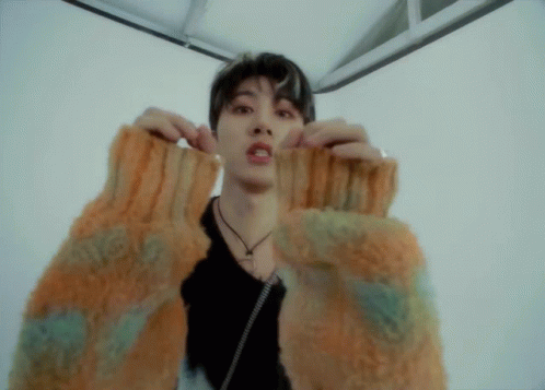 A Young Man Wearing A Sweater With A Necklace Around His Neck Looks At The Camera GIF