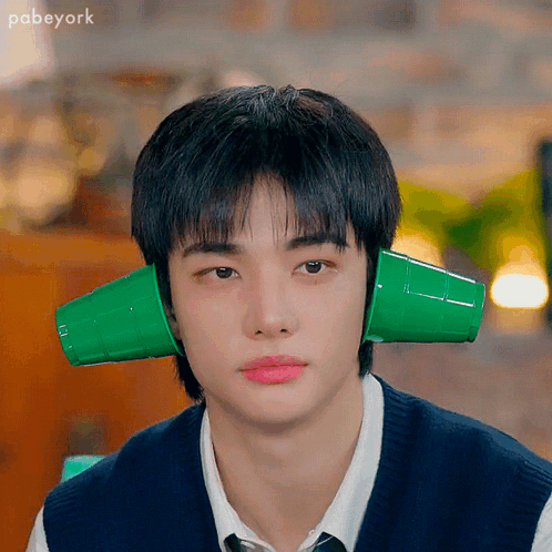 A Young Man Wearing A Vest And Tie Has Two Green Cups On His Ears . GIF