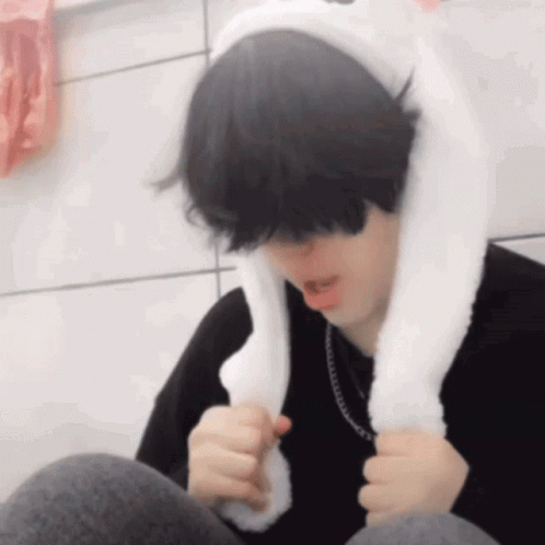 A Young Man Wearing A White Bunny Ear Hat Is Sitting Down . GIF